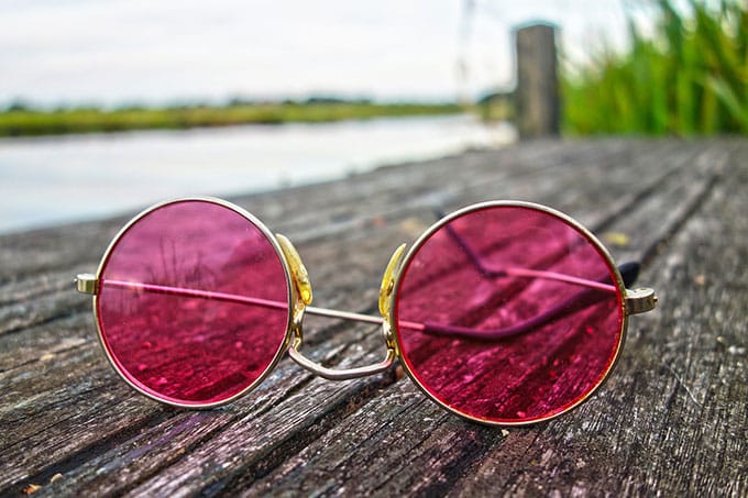 a pink colored eyeglass on a wooden top signifying that the wearer has a different experience when he views things putting it on. This talks of selective attention psychology.